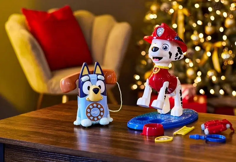 Two children's toys are placed on a wooden table in a festive setting. One toy is a blue and orange rotary-style phone shaped like a cartoon dog, and the other is a red firefighter-themed dog figure standing on a blue base with small accessories around it. In the background, there is a decorated Christmas tree with lights and ornaments, and a beige chair with a red cushion.
